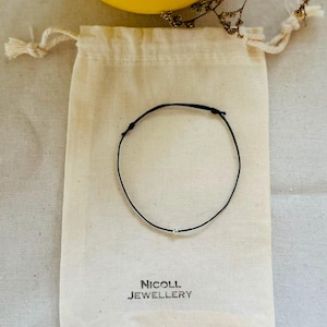 May include: A thin black cord bracelet featuring a single small silver-colored bead at the center. The bracelet is placed on a beige fabric drawstring pouch that has the text NICOLL JEWELLERY printed in black ink near the bottom edge.