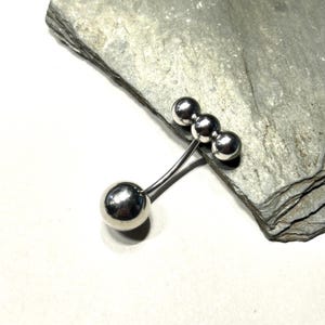 Puede incluir: Un broche plateado con un dise&ntilde;o &uacute;nico. El broche presenta cuatro formas esf&eacute;ricas: una esfera grande conectada a tres esferas m&aacute;s peque&ntilde;as por varillas met&aacute;licas finas y curvas. El broche se muestra sobre una superficie de piedra gris texturizada.