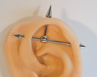 May include: A silver barbell with three spikes, one on each end and one in the middle, is shown in a pierced ear. The barbell is a popular type of jewelry for ear piercings.