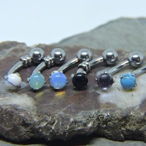 Pode incluir: Seis peças de joalheria corporal de barra curva prateadas com pedras preciosas de diferentes cores. As pedras preciosas são brancas, azul claro, azul escuro, pretas, roxas e turquesa.