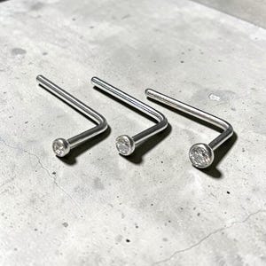 Könnte beinhalten: Drei L-förmige Nasenstecker aus poliertem, silberfarbenem Metall. Jeder Stecker verfügt über einen kleinen, runden, klaren facettierten Edelstein am Ende des geraden Stifts, was ein einfaches und funktionales Design für Gesichtsschmuck ergibt.