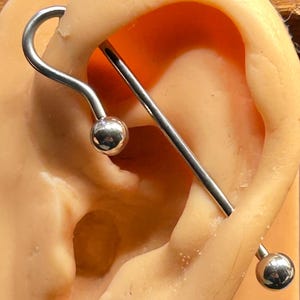 May include: A silver industrial barbell earring with two round, shiny balls. The earring is curved on one end and straight on the other, designed to fit through multiple ear piercings. The earring is shown in a tan ear model.