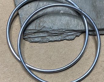 May include: Two silver-colored metal hoop earrings. The hoops are circular and have a smooth, shiny surface. They are resting on a gray, textured surface, with a brown surface in the background. The earrings are a simple, minimalist design.