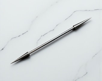 Puede incluir: Una barra para corbata de color plateado con una barra central recta y delgada y extremos cónicos puntiagudos. La barra para corbata de metal está colocada sobre un fondo de mármol blanco. Este accesorio está diseñado para mantener una corbata en su lugar.