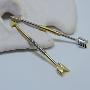 Könnte beinhalten: Zwei industrielle Barbell-Ohrringe mit pfeilförmigen Enden. Ein Ohrring hat einen goldfarbenen Pfeil und eine goldfarbene gewickelte Mitte, während der andere einen silberfarbenen Pfeil und eine silberfarbene gewickelte Mitte hat. Die Stäbe sind silberfarben.