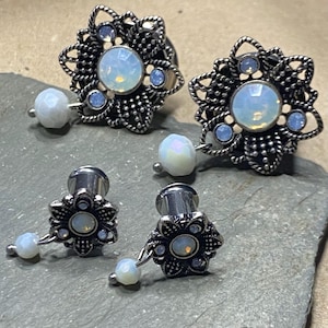 Op de afbeelding: Vier sieraden. Twee zijn groter, met een bloemmotief en een centrale opalescente steen, omringd door kleinere blauwe stenen. De andere twee zijn kleiner, met een vergelijkbaar ontwerp en een bungelende opalescente kraal. Allemaal zilverkleurig.
