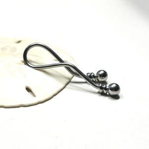 May include: A silver-toned metal body jewelry piece featuring a curved, twisted bar design that forms an elongated loop. The ends of the bar are finished with two polished spherical beads, each secured by a small, coiled metal detail at the base.