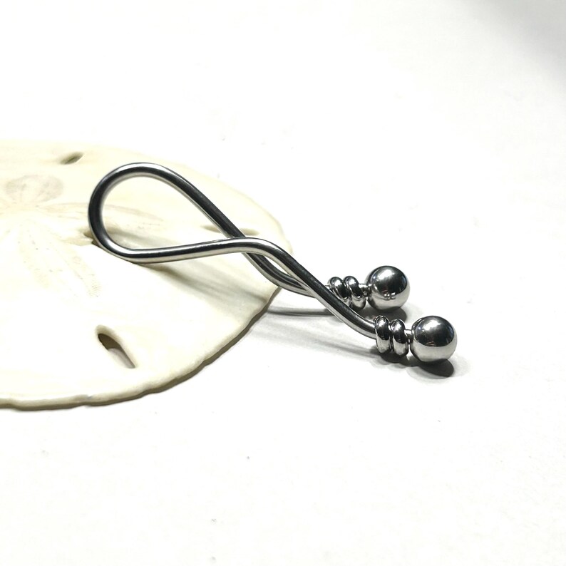 May include: A silver-toned metal body jewelry piece featuring a curved, twisted bar design that forms an elongated loop. The ends of the bar are finished with two polished spherical beads, each secured by a small, coiled metal detail at the base.