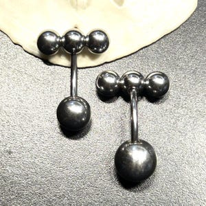 Puede incluir: Dos piercings de barra de color plateado. Cada barra presenta tres peque&ntilde;as esferas conectadas en fila en la parte superior, con una esfera m&aacute;s grande en la parte inferior. Los piercings se muestran sobre una superficie texturizada.