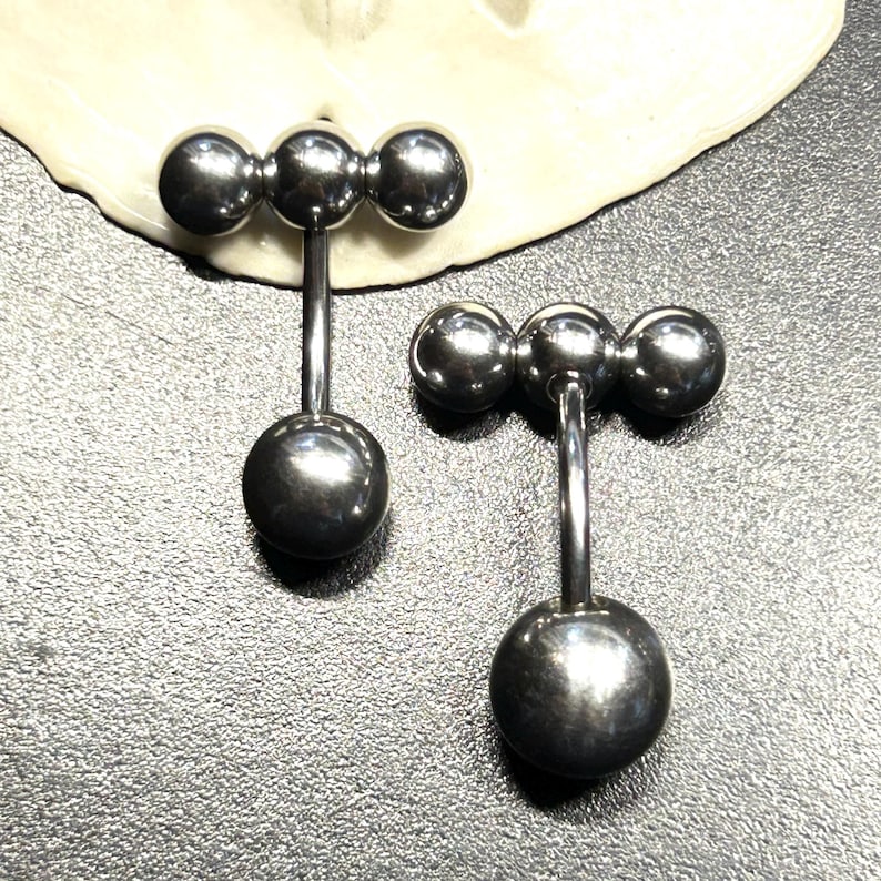 Puede incluir: Dos piercings de barra de color plateado. Cada barra presenta tres peque&ntilde;as esferas conectadas en fila en la parte superior, con una esfera m&aacute;s grande en la parte inferior. Los piercings se muestran sobre una superficie texturizada.