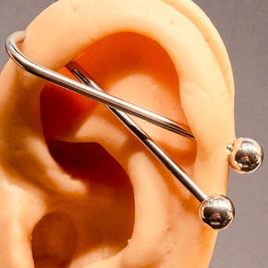 May include: A silver industrial barbell earring with two round ball ends. The earring is shown on a light-colored ear model, with the barbell extending across the upper ear. The jewelry is made of shiny, polished metal.