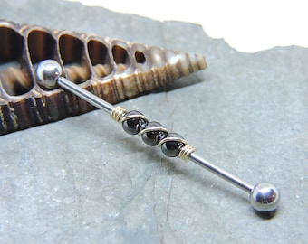 May include: A silver barbell with three black beads wrapped in gold wire. The barbell is lying on a gray stone surface.