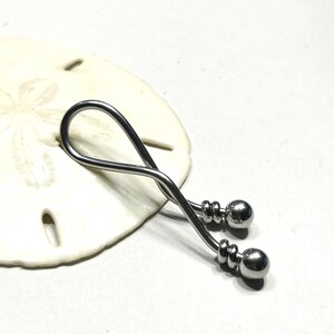 May include: A silver-toned metal body jewelry piece featuring a curved, twisted bar design that forms a loop. The ends of the bar are finished with two polished spherical beads, each secured by a small, grooved metal ring detail near the tip.