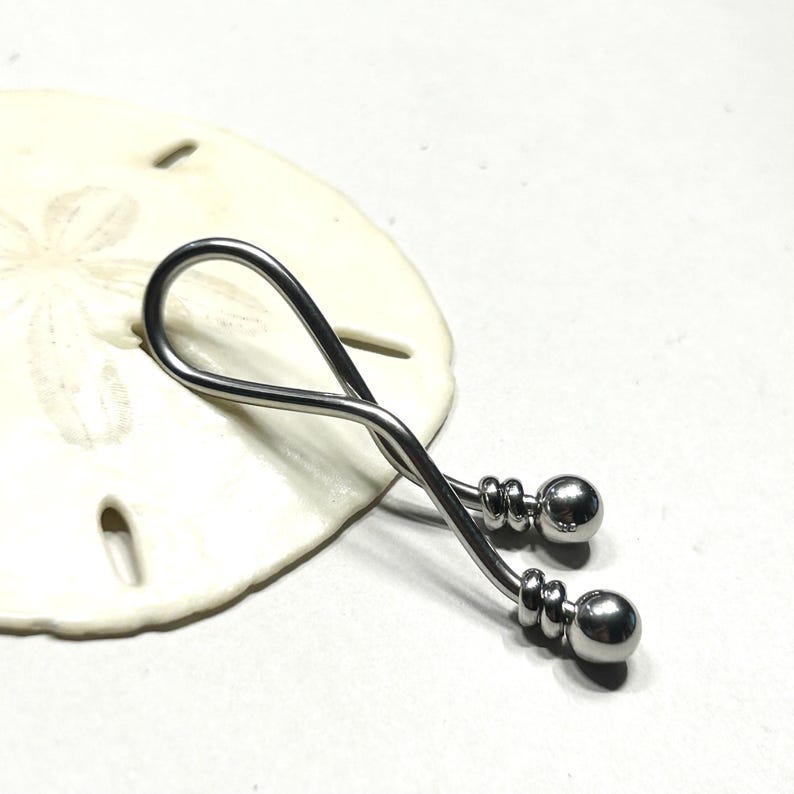 May include: A silver-toned metal body jewelry piece featuring a curved, twisted bar design that forms a loop. The ends of the bar are finished with two polished spherical beads, each secured by a small, grooved metal ring detail near the tip.