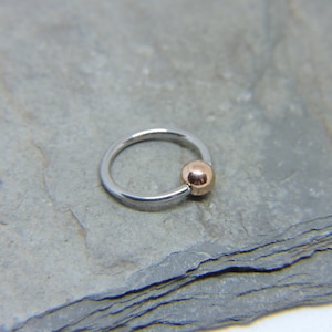 May include: A silver ring with a rose gold ball on the end. The ring is on a gray background.