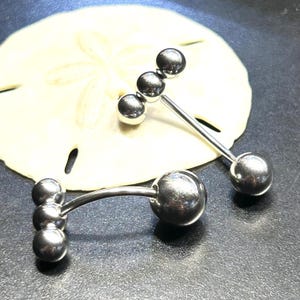 Puede incluir: Dos piercings de lengua tipo barbell plateados. Cada uno tiene una bola grande en un extremo y tres bolas m&aacute;s peque&ntilde;as en el otro. Los piercings se muestran sobre un d&oacute;lar de arena sobre un fondo oscuro.