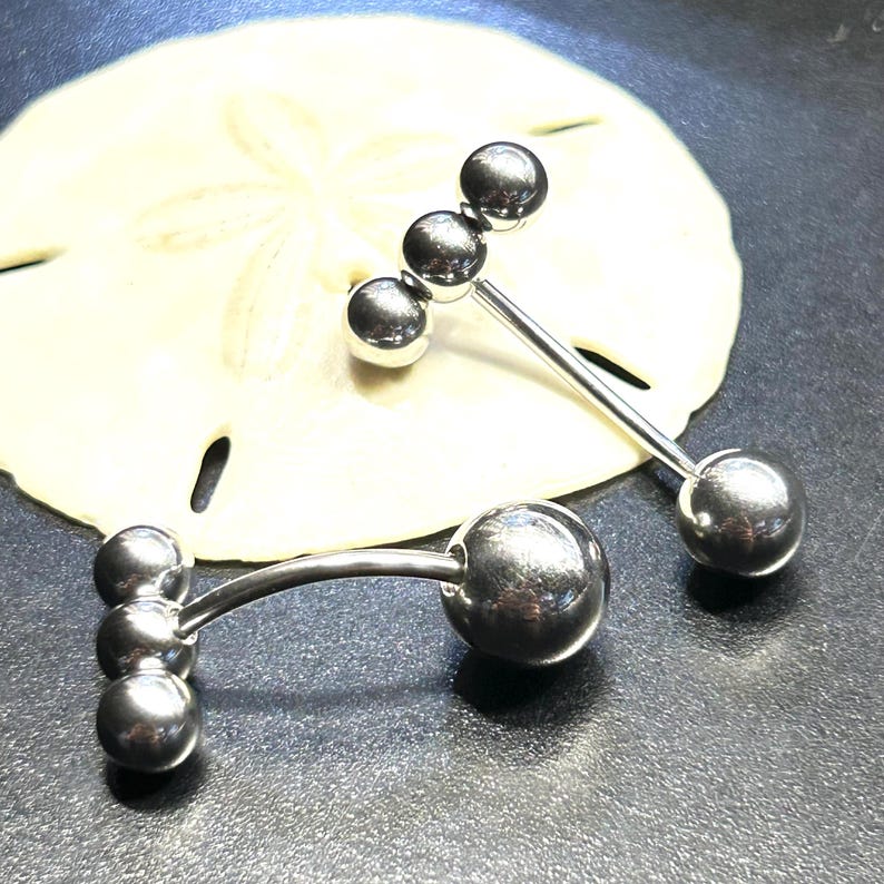 Puede incluir: Dos piercings de lengua tipo barbell plateados. Cada uno tiene una bola grande en un extremo y tres bolas m&aacute;s peque&ntilde;as en el otro. Los piercings se muestran sobre un d&oacute;lar de arena sobre un fondo oscuro.