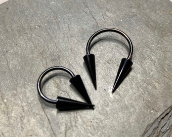 May include: Two black metal circular hoop earrings with pointed ends. The earrings are on a gray background.