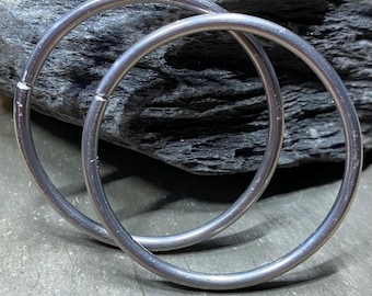 May include: Two silver hoop earrings, one slightly larger than the other, are displayed on a gray textured surface.