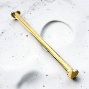 May include: A gold-toned industrial barbell body jewelry piece featuring a straight metal bar with a small heart-shaped end piece on each side. The smooth, polished surface of the metal reflects light, highlighting the simple, linear design of this accessory.