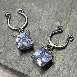 May include: Two silver-colored nipple rings with square, clear, sparkling cubic zirconia pendants. The rings have a curved design with a small loop at the top and a hook at the bottom.