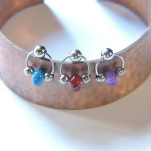 Könnte beinhalten: Drei silberne Creolen mit bunten Perlen. Die Creolen sind aus Metall und haben eine kreisförmige Form. Die Perlen sind rund und in den Farben Blau, Rot und Lila.