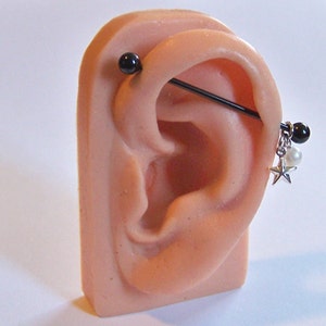 Peut inclure: Boucle d'oreille industrielle noire avec un charme pendantif en forme d'étoile, de perle et de perle noire. La boucle d'oreille est présentée sur un modèle d'oreille rose, mettant en valeur le design et l'ajustement du bijou.
