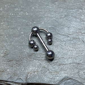 Puede incluir: Barbell de plata con una parte superior curva y múltiples bolas. La barra tiene una sola bola en la parte inferior y tres bolas en la parte superior curva.