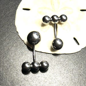 Puede incluir: Dos piercings para el ombligo de color plateado. Uno tiene una bola grande en la parte superior y tres bolas m&aacute;s peque&ntilde;as en la parte inferior. El otro tiene tres bolas peque&ntilde;as en la parte superior y una bola grande en la parte inferior. Los anillos se muestran sobre una superficie blanca y texturizada.
