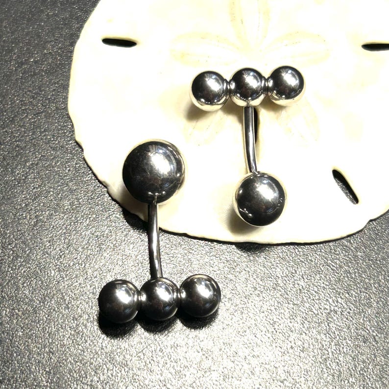 Puede incluir: Dos piercings para el ombligo de color plateado. Uno tiene una bola grande en la parte superior y tres bolas m&aacute;s peque&ntilde;as en la parte inferior. El otro tiene tres bolas peque&ntilde;as en la parte superior y una bola grande en la parte inferior. Los anillos se muestran sobre una superficie blanca y texturizada.