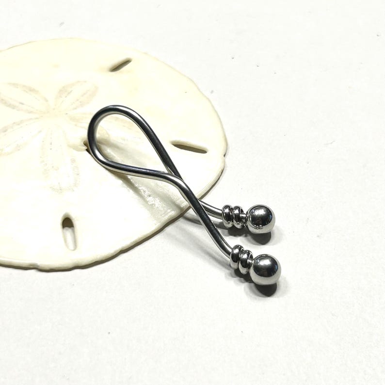 May include: A silver-toned metal body jewelry piece featuring a curved, twisted bar design that forms a loop at one end. The two ends of the bar are finished with polished spherical beads, each accented by a small grooved ring detail near the base.