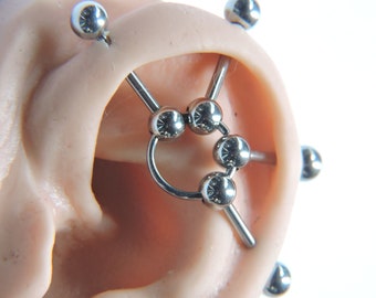 May include: A silver industrial barbell with a circular design and multiple balls. The barbell is inserted into a simulated ear piercing.