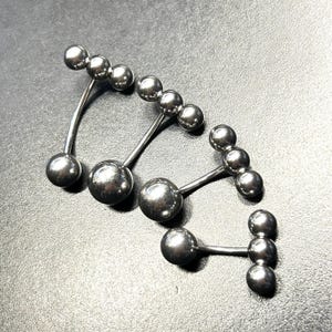Puede incluir: Cuatro piezas de joyer&iacute;a corporal plateadas. Cada pieza tiene una barra curva o recta con m&uacute;ltiples cuentas esf&eacute;ricas. La joyer&iacute;a est&aacute; hecha de un material met&aacute;lico brillante y se muestra sobre una superficie oscura.