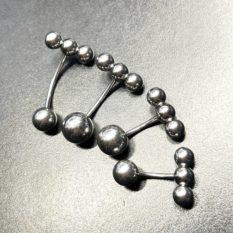Puede incluir: Cuatro piezas de joyer&iacute;a corporal plateadas. Cada pieza tiene una barra curva o recta con m&uacute;ltiples cuentas esf&eacute;ricas. La joyer&iacute;a est&aacute; hecha de un material met&aacute;lico brillante y se muestra sobre una superficie oscura.