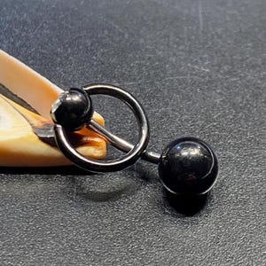 Könnte beinhalten: Ein schwarzer Barbell-Körperpiercing mit einem kreisförmigen Ring und zwei schwarzen kugelförmigen Enden. Ein Ende hat einen kleinen, klaren Edelstein. Der Schmuck ist vor einem dunklen Hintergrund platziert.