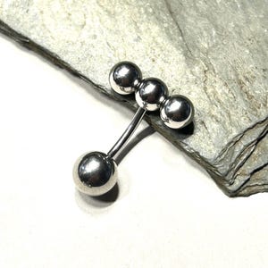 Puede incluir: Un piercing de barra plateada con cuatro bolas esf&eacute;ricas. La barra tiene un dise&ntilde;o curvo con tres bolas agrupadas y una bola en el extremo opuesto. La joya se exhibe sobre una superficie de piedra gris.