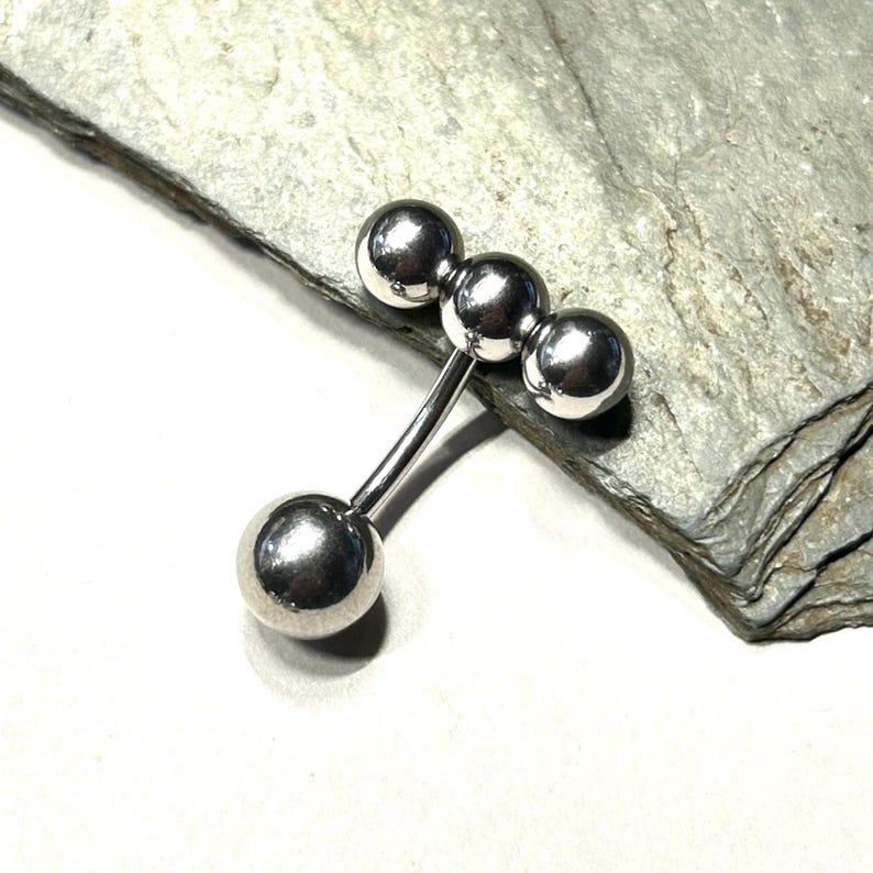 Puede incluir: Un piercing de barra plateada con cuatro bolas esf&eacute;ricas. La barra tiene un dise&ntilde;o curvo con tres bolas agrupadas y una bola en el extremo opuesto. La joya se exhibe sobre una superficie de piedra gris.
