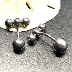 Puede incluir: Dos piercings para el ombligo de color plateado. Cada uno tiene una bola grande y redonda en la parte inferior y tres bolas m&aacute;s peque&ntilde;as en la parte superior, conectadas por una barra curva. Las joyas se muestran sobre una superficie oscura.