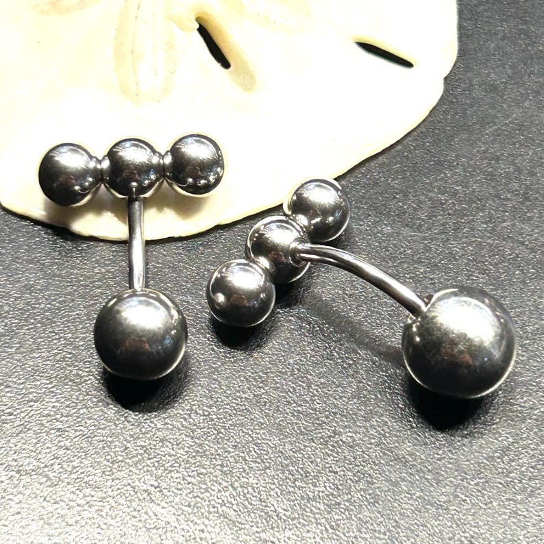 Puede incluir: Dos piercings para el ombligo de color plateado. Cada uno tiene una bola grande y redonda en la parte inferior y tres bolas m&aacute;s peque&ntilde;as en la parte superior, conectadas por una barra curva. Las joyas se muestran sobre una superficie oscura.