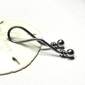 May include: A silver-toned metal body jewelry piece featuring a curved, twisted bar design. The ends of the bar are finished with polished spherical beads, each accented by two small, grooved rings positioned just below the ball terminals.