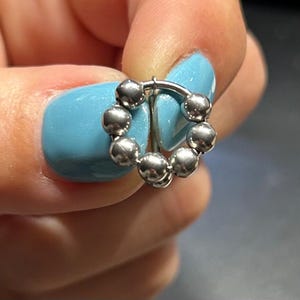 May include: A silver-colored, circular earring with a series of small, round beads forming a decorative ring. The earring is held between fingers with light blue nail polish.
