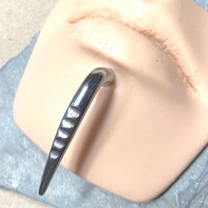 May include: A silver-toned curved spike piercing jewelry piece featuring a series of five small, rounded indentations along the outer edge. The metal has a polished, reflective finish and is shown inserted into a light-toned synthetic skin model.