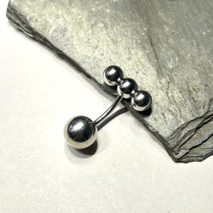 Puede incluir: Una joya de piercing corporal plateada, estilo barra. Presenta tres peque&ntilde;as esferas conectadas a una barra curva, con una esfera m&aacute;s grande en el extremo opuesto. La pieza se exhibe sobre una superficie de piedra gris.
