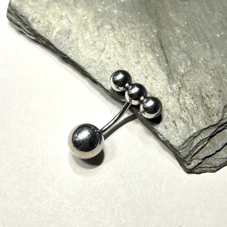 Puede incluir: Una joya de piercing corporal plateada, estilo barra. Presenta tres peque&ntilde;as esferas conectadas a una barra curva, con una esfera m&aacute;s grande en el extremo opuesto. La pieza se exhibe sobre una superficie de piedra gris.