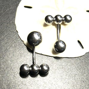 Puede incluir: Dos piercings para el ombligo de color plateado. Un anillo tiene una bola grande en la parte superior y tres bolas m&aacute;s peque&ntilde;as en la parte inferior. El otro anillo tiene tres bolas en la parte superior y una sola bola en la parte inferior. Los anillos se muestran sobre una superficie blanca y texturizada.
