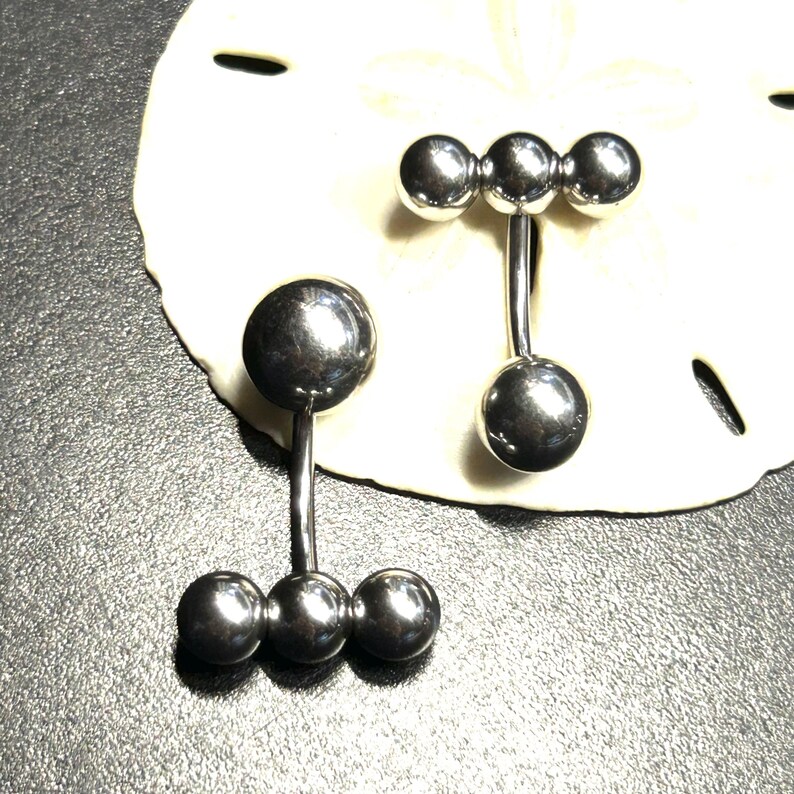 Puede incluir: Dos piercings para el ombligo de color plateado. Un anillo tiene una bola grande en la parte superior y tres bolas m&aacute;s peque&ntilde;as en la parte inferior. El otro anillo tiene tres bolas en la parte superior y una sola bola en la parte inferior. Los anillos se muestran sobre una superficie blanca y texturizada.