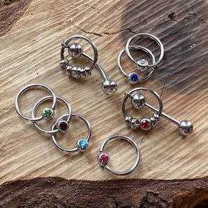 May include: Assortment of silver body jewelry with colorful gemstones. The jewelry includes captive bead rings, a hinged segment ring, and a barbell with a rotating ring. The gemstones are red, blue, green, and clear.