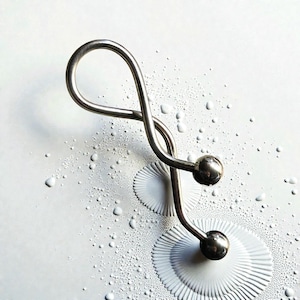 May include: A silver-colored metal object with a looped top and two spherical ends. The object is on a white surface with small, scattered droplets and two circular, textured patterns.