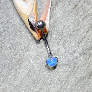 Pode incluir: Um piercing corporal de prata com uma gema de opala azul claro. O piercing é mostrado contra um fundo cinza.