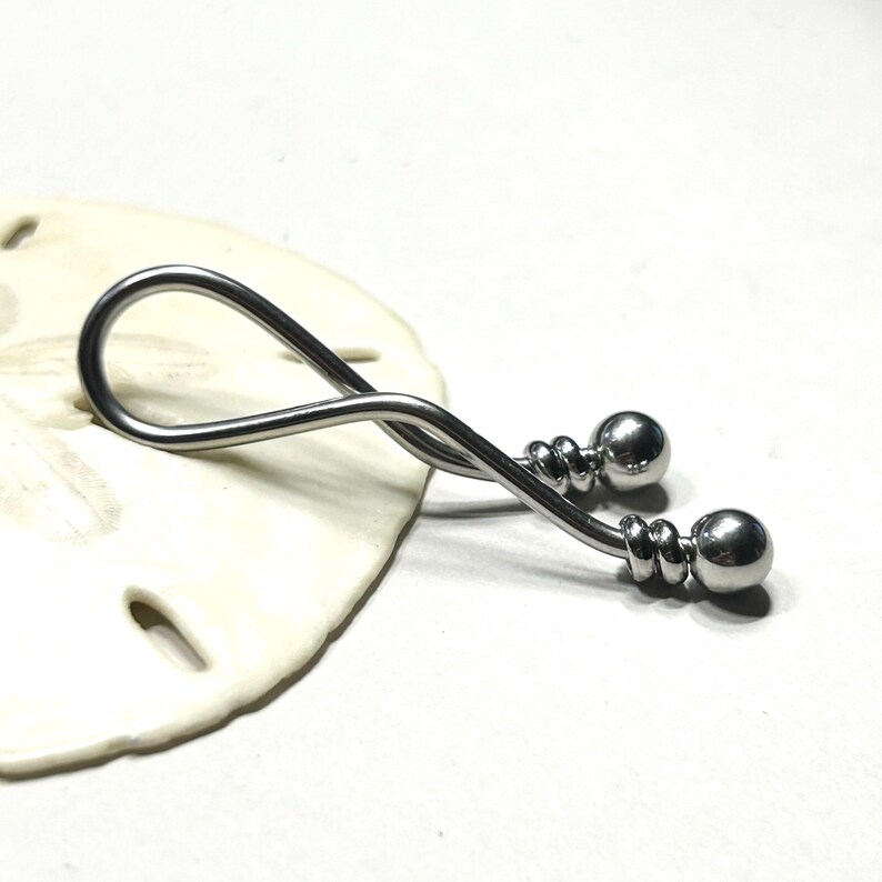 May include: A silver-toned metal body jewelry piece featuring a curved, twisted bar design. The ends of the bar are finished with polished spherical beads, each accented by two small, grooved rings positioned just below the ball terminals.